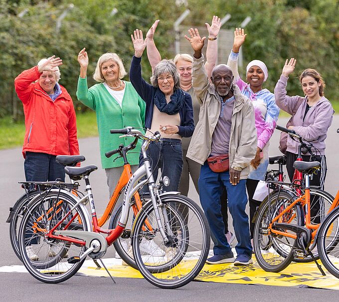 Vielfältige Gruppe Eine Gruppe steht auf einer asphaltierten Fläche und winkt freundlich in die Kamera. Vor ihnen stehen drei orangefarbene Fahrräder. Die Gruppe wirkt fröhlich und gemeinschaftlich, im Hintergrund ist Grünbewuchs und ein Zaun zu sehen.