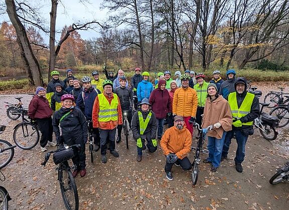Gruppenfoto von gut gelaunten Radfahrenden in wetterfester Kleidung, die mit ihren Fahrrädern auf einem laubbedeckten Waldweg stehen. Viele tragen Fahrradhelme und einige gelbe Warnwesten. Im Hintergrund sind herbstliche Bäume und ein bewölkter Himmel zu sehen.